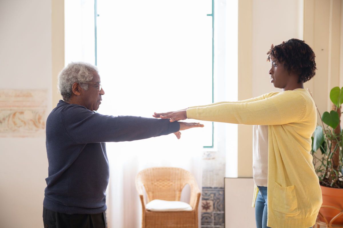 A carer and an older man stand facing each other by a bright window doing an arm-raising exercise at home