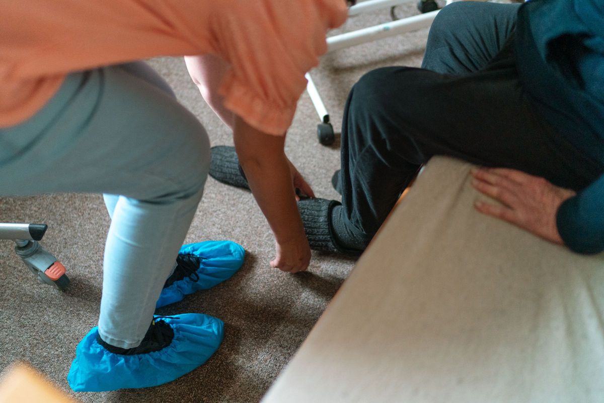 A carer kneels to help an older man with his socks beside his bed, with a walking frame nearby