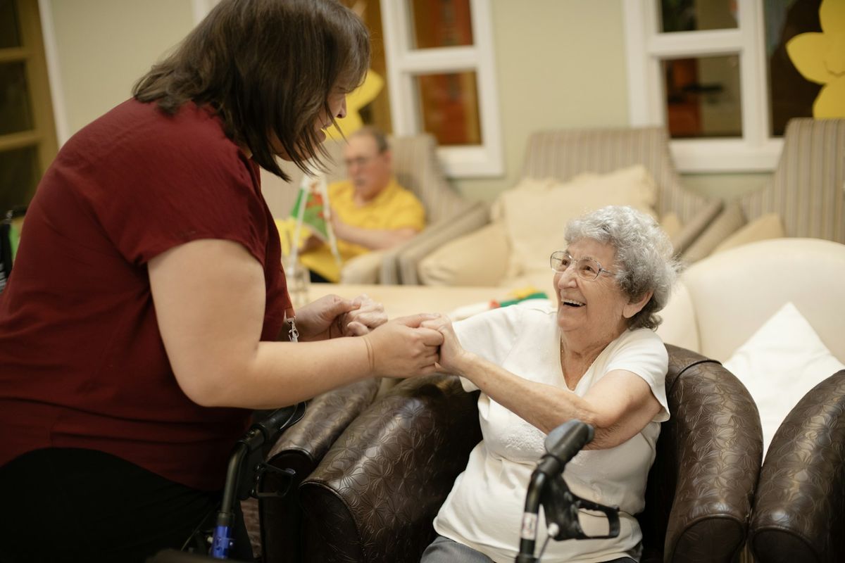 A smiling older woman in an armchair holding hands with a younger carer, with a walking frame nearby