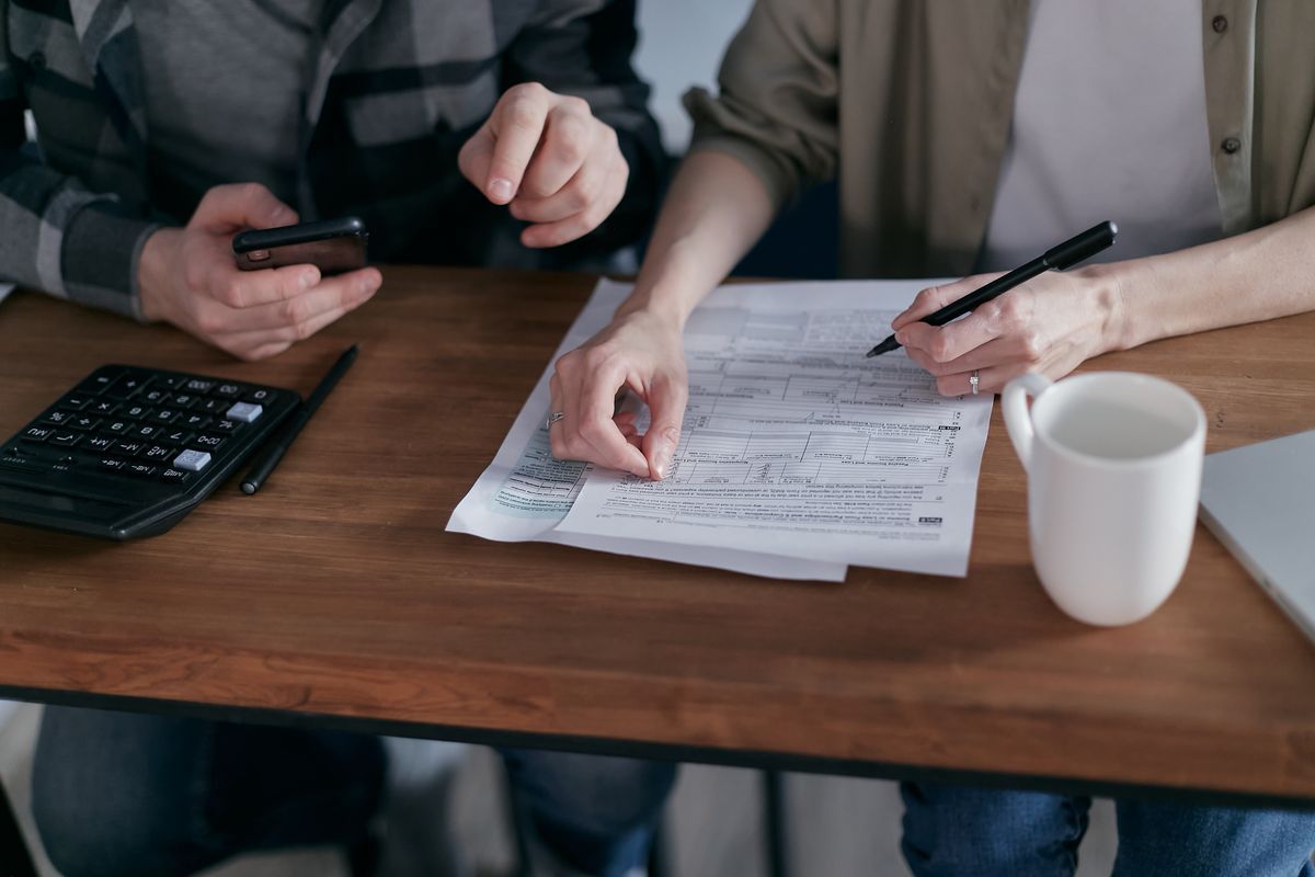 Two people at a wooden table going through paperwork with a calculator, phone and mug of tea