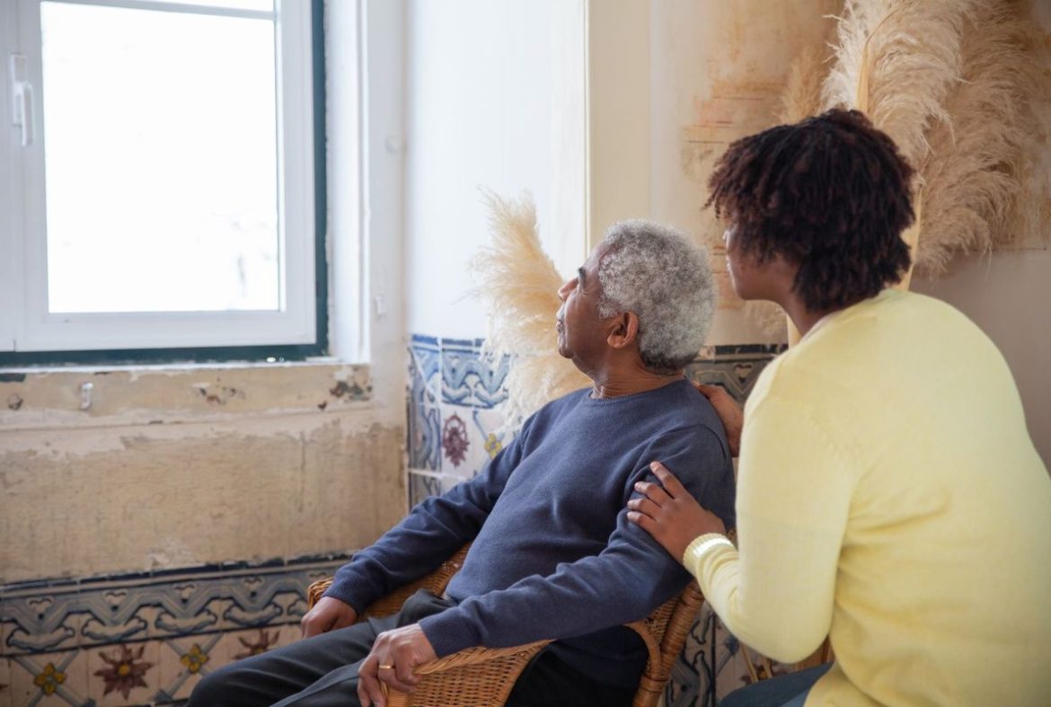 A younger woman resting a hand on an older man's shoulder as they look out a window at home