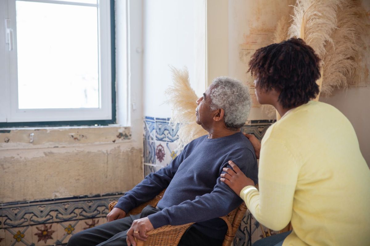 A younger woman resting a hand on an older manâ€™s shoulder as they look out a window at home