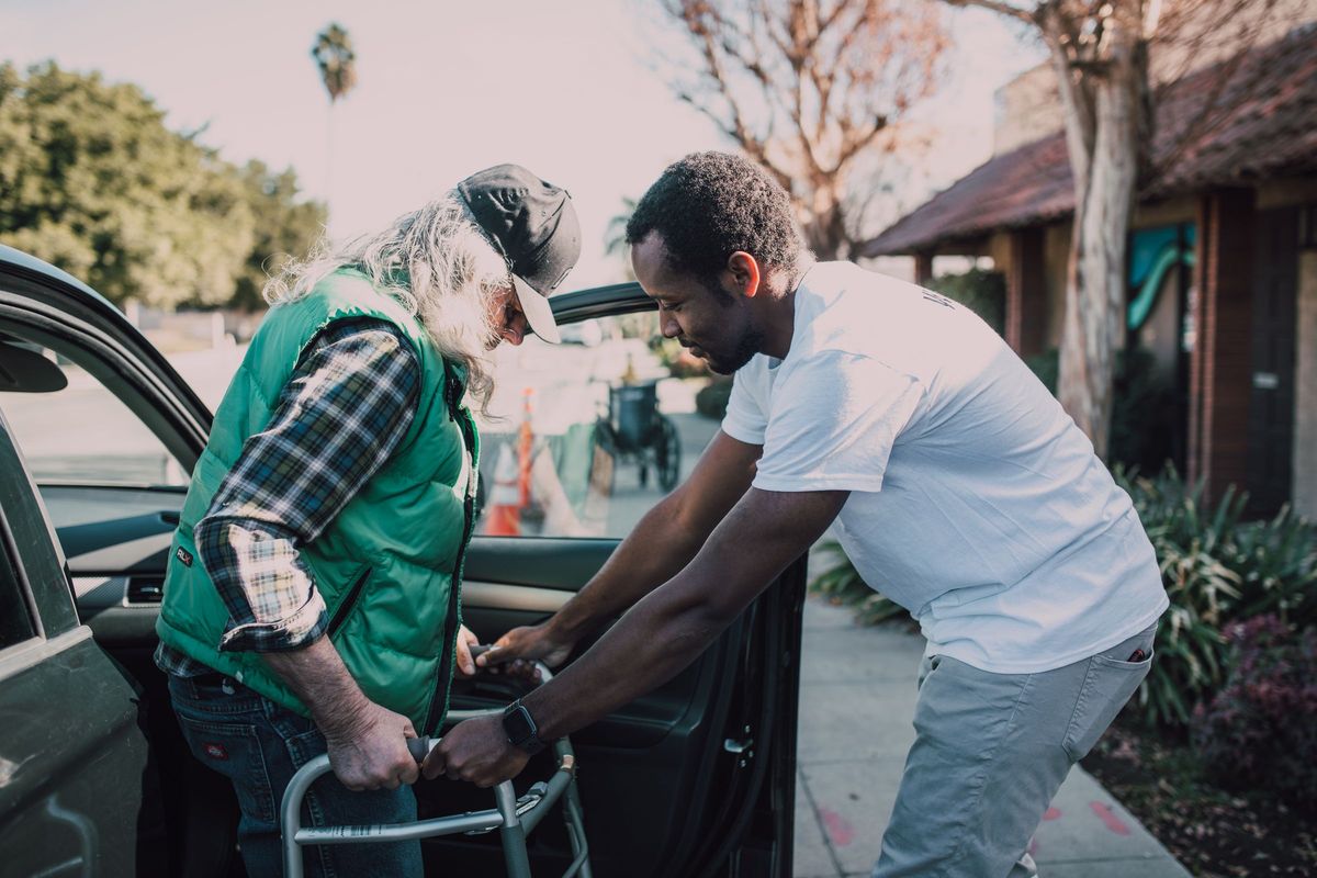 A younger man steadies an older man's walking frame as he gets out of a car