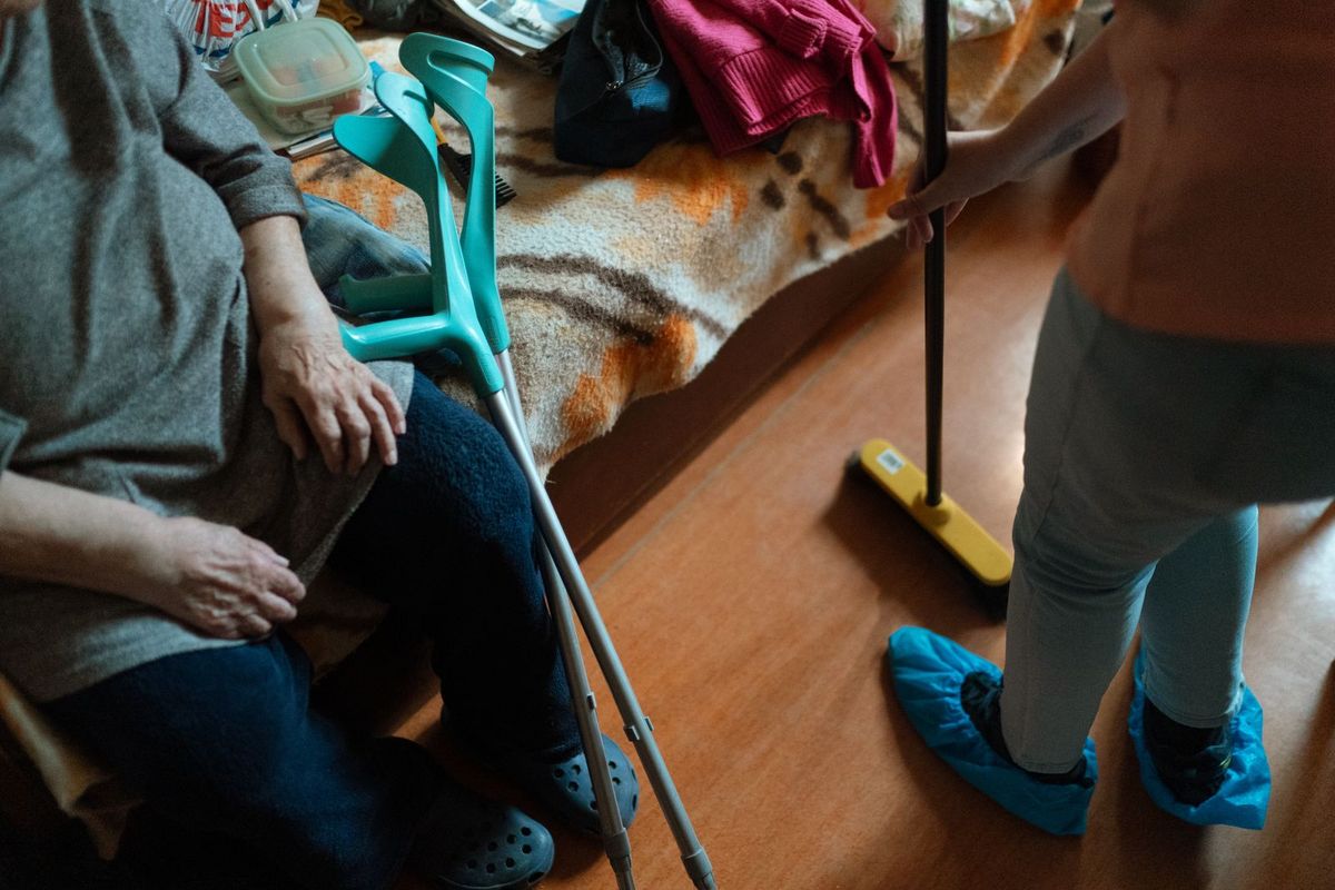 A person in blue shoe covers sweeping a wooden floor beside an older person sitting with crutches nearby