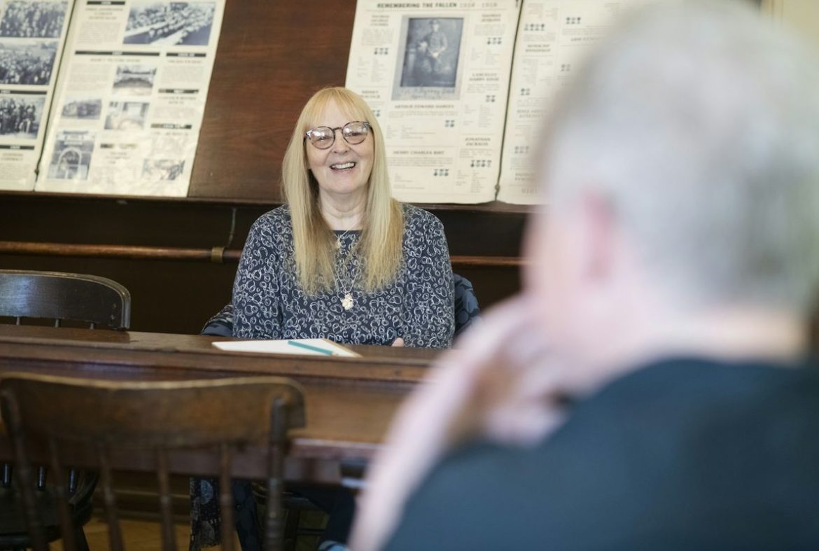 An older woman with glasses smiling across a wooden table during a conversation
