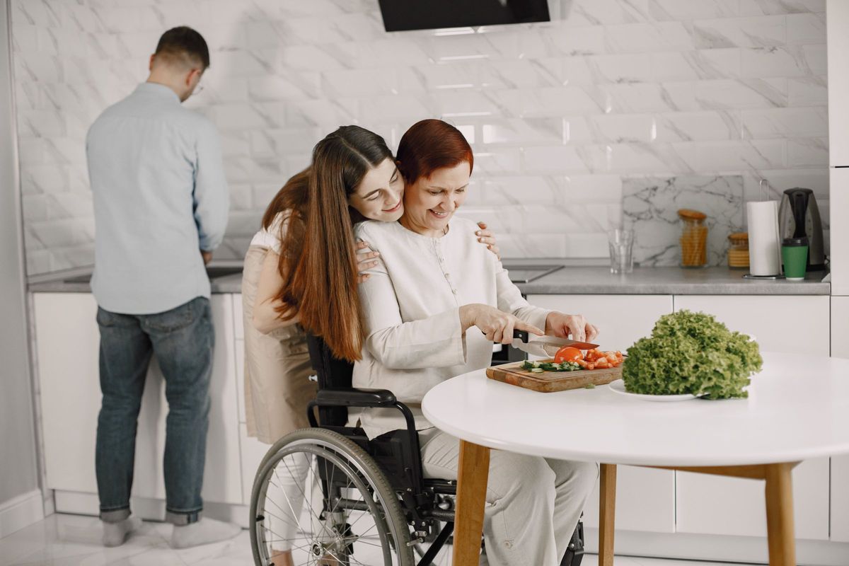 An older woman in a wheelchair preparing vegetables at a kitchen table while her daughter hugs her