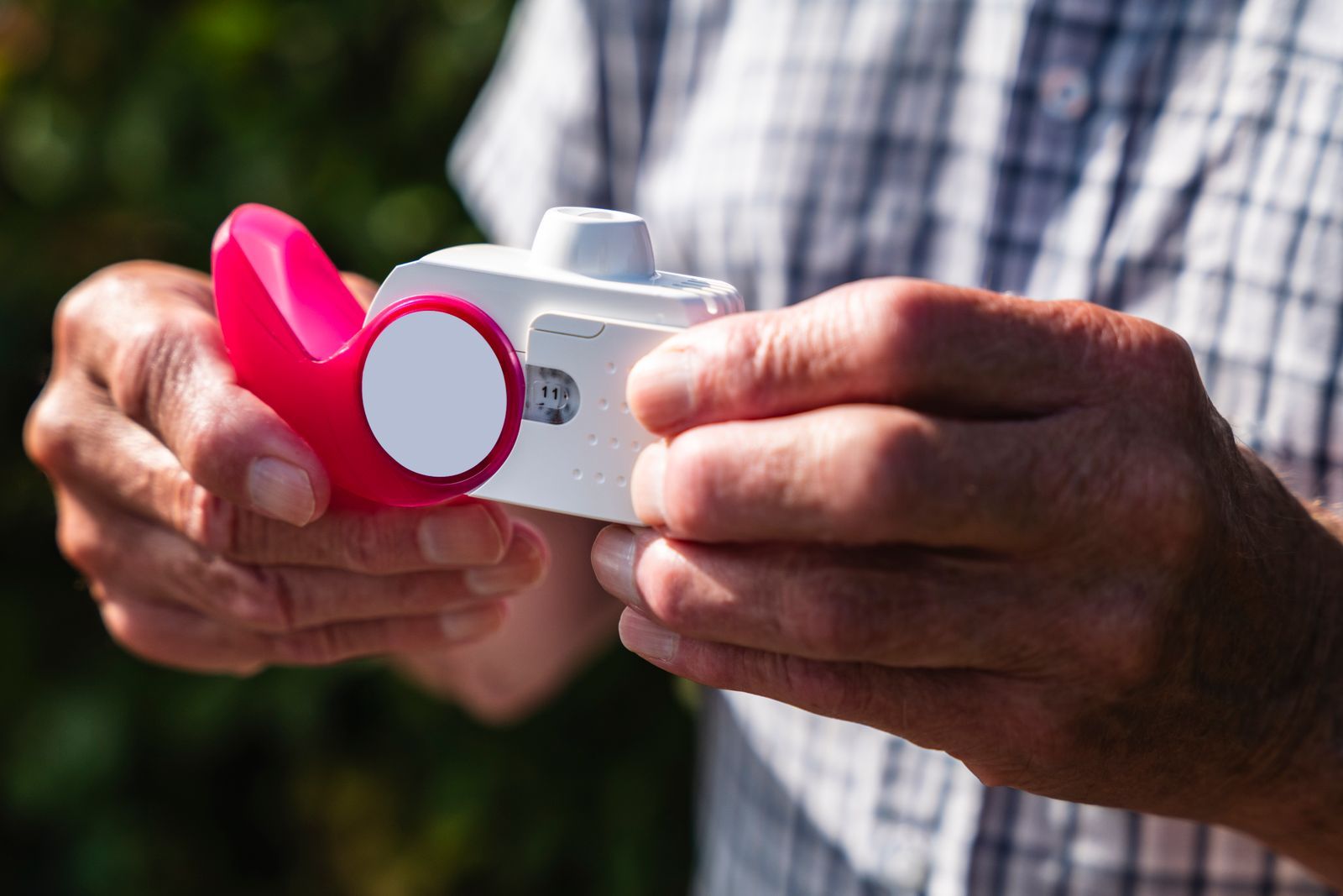 An older person's hands hold a white and pink inhaler in front of a checked shirt, outdoors in soft sunlight
