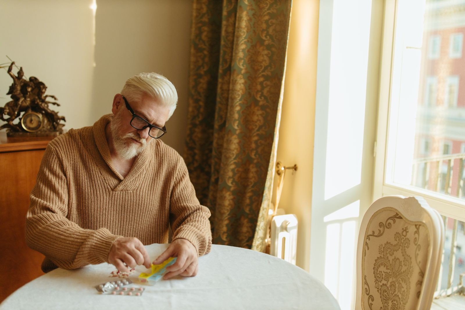An older man with grey hair and glasses sorts tablets into a weekly pill organiser at a table by a window at home