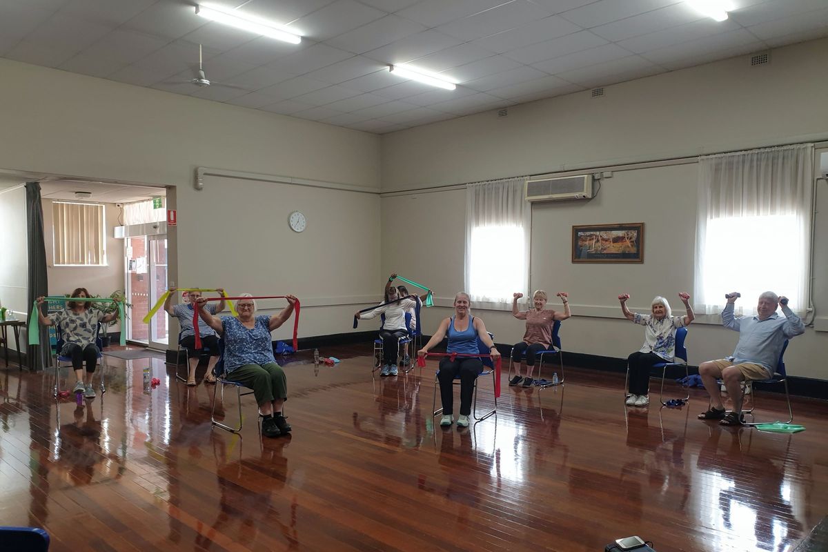 Older people doing a seated strength exercise class with resistance bands and hand weights in a community hall