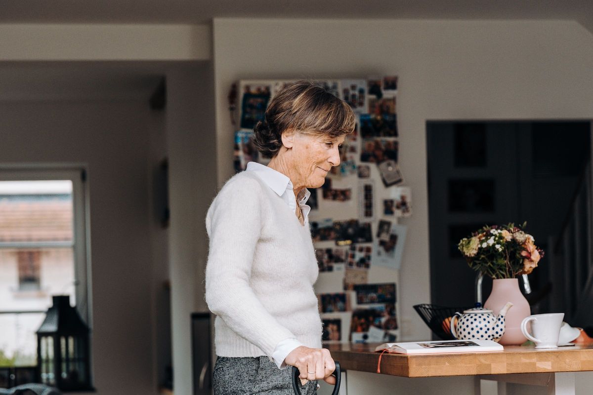 An older woman smiling as she walks through her kitchen holding the handle of a walking aid