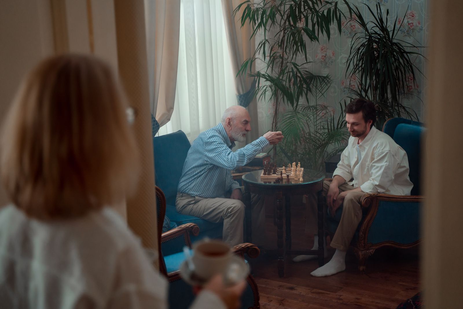 An adult son plays chess with his older father in a living room at home while a woman holding a cup of tea looks on
