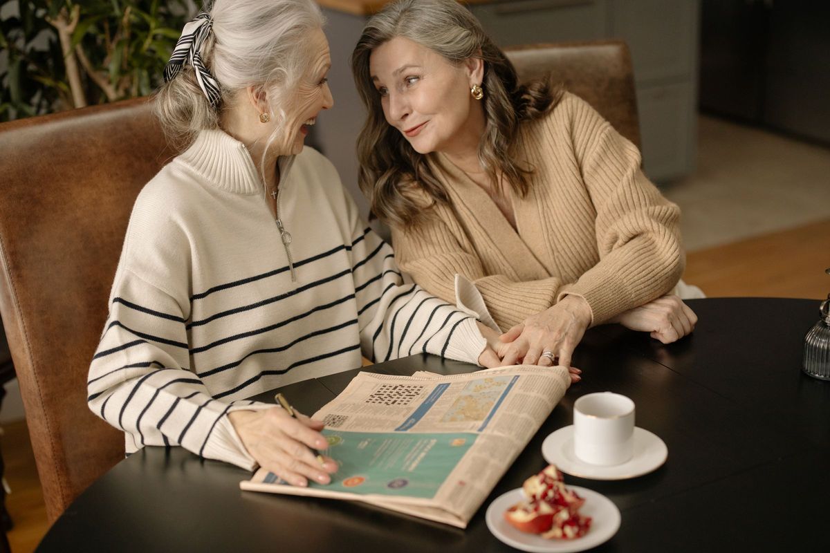 Two older women laughing together over a newspaper crossword and a cup of tea at home