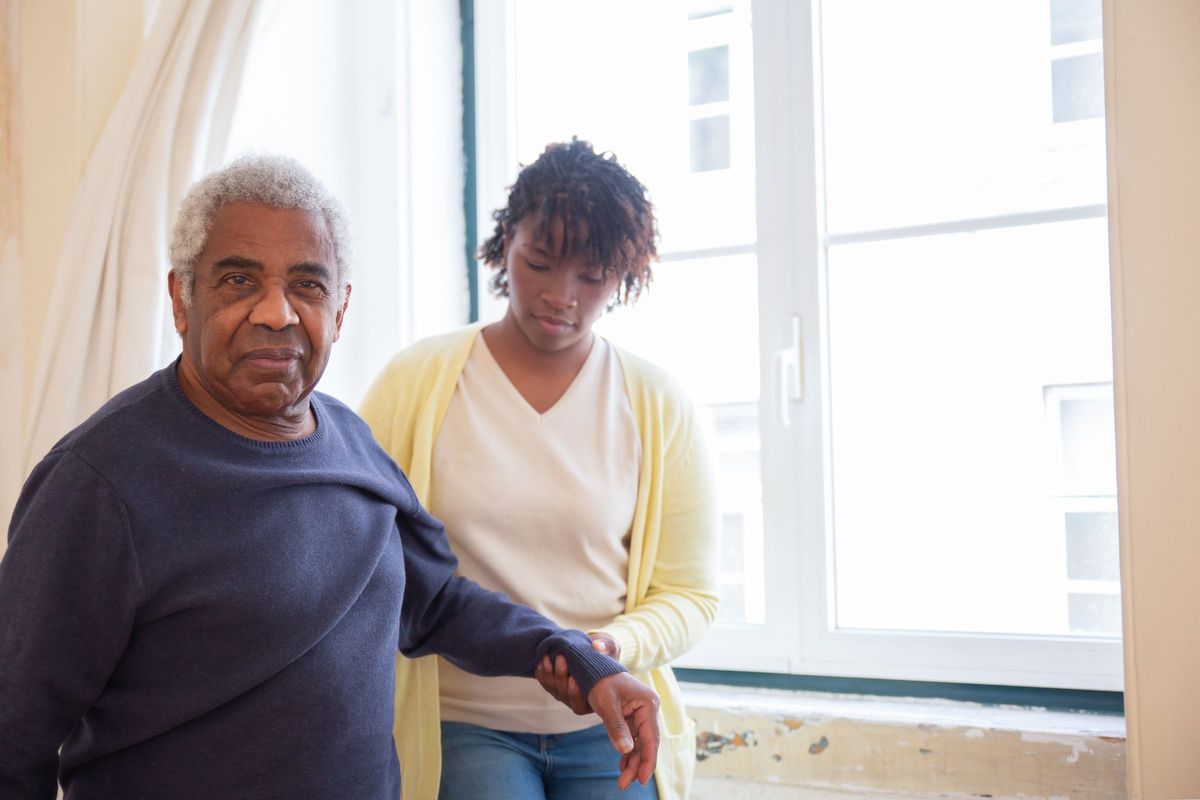 A younger woman steadying an older man's arm as he walks beside a bright window at home
