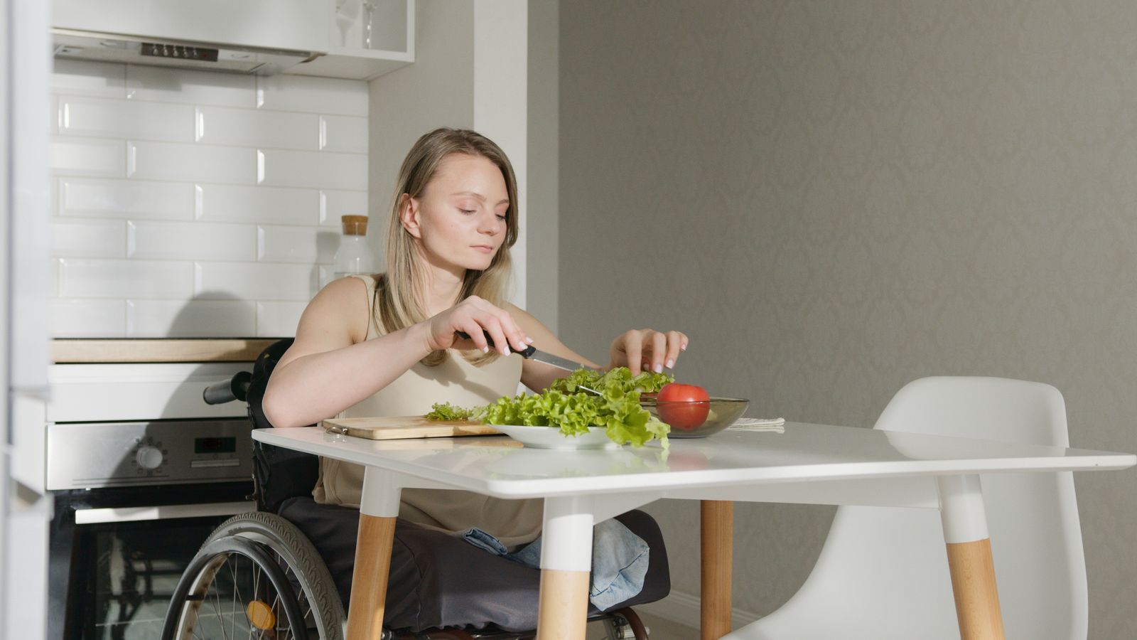 A young woman in a wheelchair chops lettuce and a tomato at a white table in her kitchen at home